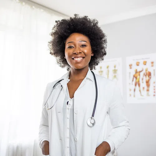 Smiling female doctor in white coat with stethoscope standing in bright medical office with anatomy charts.