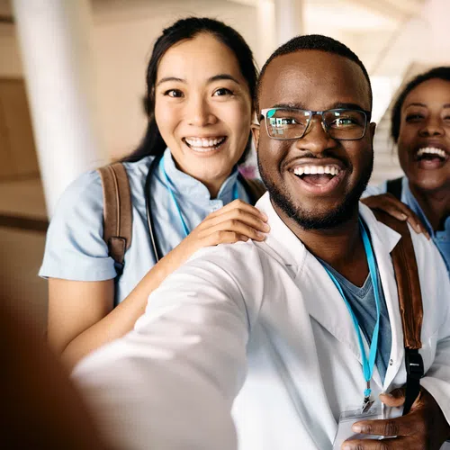 Diverse group of smiling medical students and doctor taking a cheerful selfie indoors, wearing scrubs and lab coats.