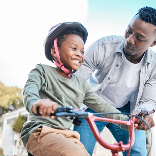 Smiling father helps young boy wearing helmet ride bicycle outdoors on a sunny day.