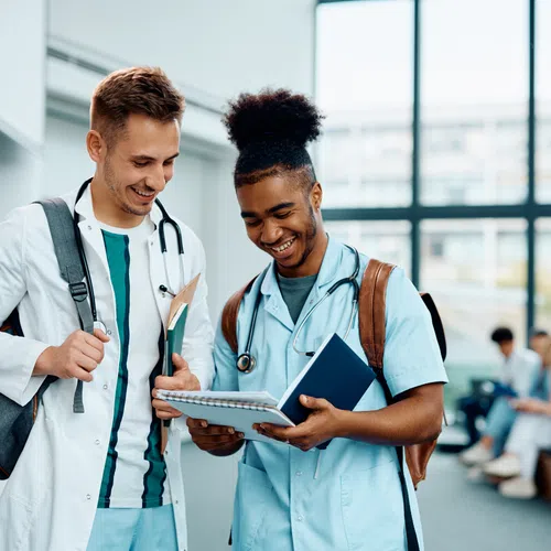 Two medical students in scrubs and lab coat smiling while reviewing notes together in a bright campus lobby.
