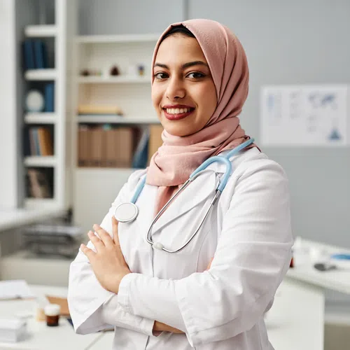 Young female doctor wearing a pink hijab and white coat with stethoscope smiling in a bright medical office.