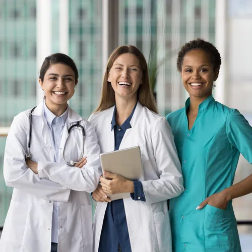 Three female healthcare professionals smiling, two in white coats and one in scrubs, standing in a medical facility.
