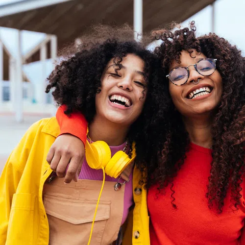 Two joyful young women with curly hair smiling outdoors, wearing bright colorful clothing and headphones