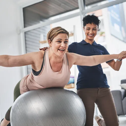 Woman balancing on exercise ball with arms extended guided by smiling personal trainer indoors