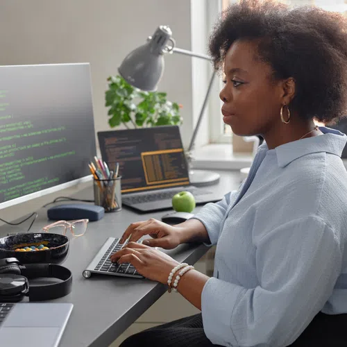 Woman working on coding at a computer desk with multiple screens and office supplies in a modern workspace.