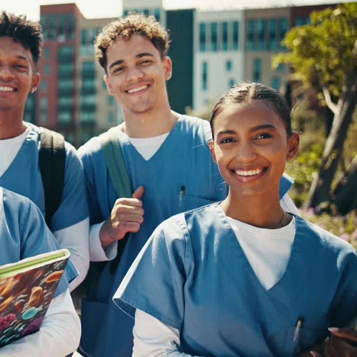 Group of five diverse nursing students in blue scrubs smiling outdoors with books and backpacks in urban setting.