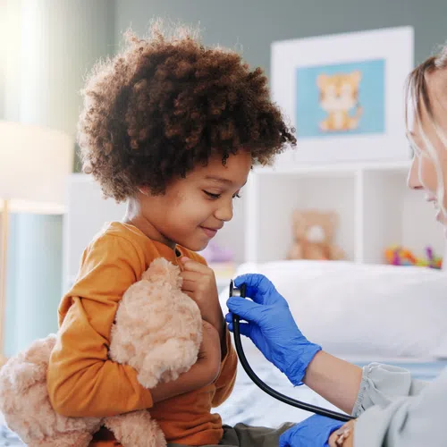Doctor using stethoscope to check a smiling child's heartbeat while the child holds a teddy bear in a cozy room.