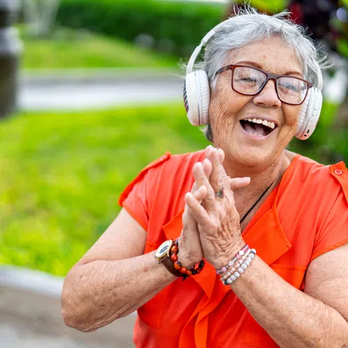 Joyful elderly woman in orange shirt wearing headphones and clapping hands outdoors in a garden setting