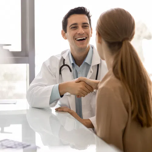 Smiling male doctor shaking hands with female patient in bright medical office during consultation.