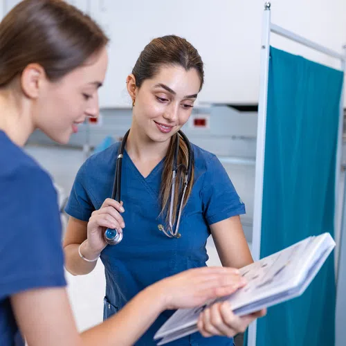 Two female medical professionals in blue scrubs review patient charts in a clinical setting with a privacy screen.