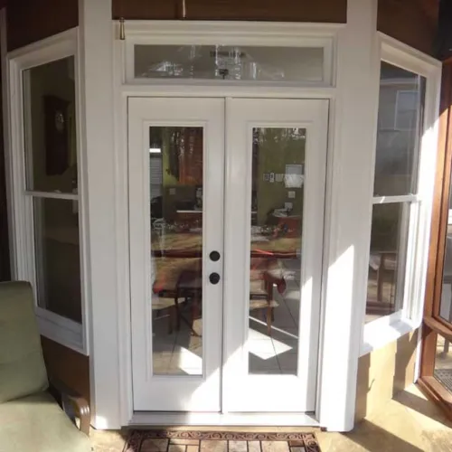White double glass doors with black handles opening to a sunlit interior patio space with green chair.