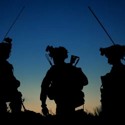 Silhouettes of three armed soldiers with gear against a twilight sky in a rugged outdoor setting