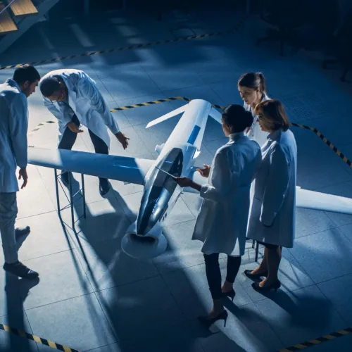 A group of engineers in lab coats examine a white drone aircraft prototype inside a marked testing area.