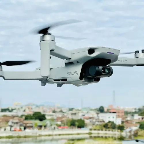 White DJI Mini 2 drone flying over an urban area with blurred buildings and cloudy sky background.