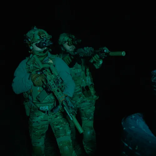 Two soldiers in night vision gear and camouflage with rifles advancing cautiously in the dark near stacked tires.