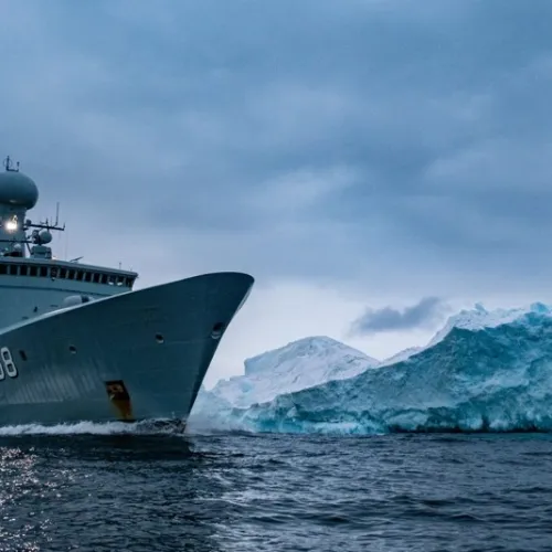 Military ship F358 sailing near a large iceberg in cold, cloudy Arctic waters at dusk.