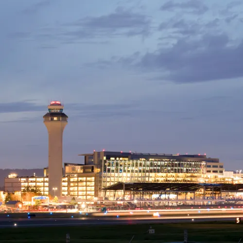 Night view of illuminated airport terminal and control tower under a cloudy sky with runway lights in foreground