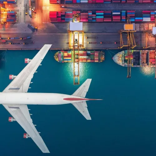 Aerial view of a large white airplane flying over a port with container cranes and cargo ships at dusk.