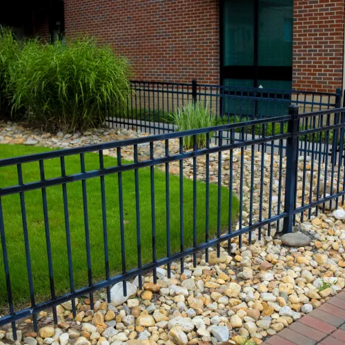 Black metal fence enclosing green grass and decorative rocks beside a brick building exterior.