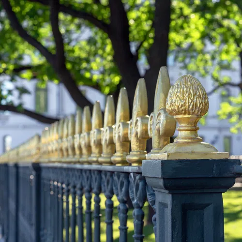 Close-up of a decorative black fence with gold accents, surrounded by lush green trees and sunlight.