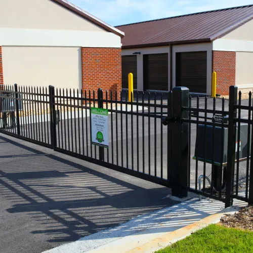 Black metal fence and gate securing a parking area outside brick and beige commercial buildings on a sunny day.
