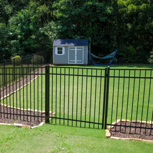 Black metal fence enclosing a grassy yard with a gray shed and hammock near a wooded area.