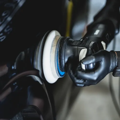 Close-up of hands using a polishing machine with white pad on a black car surface wearing black gloves.