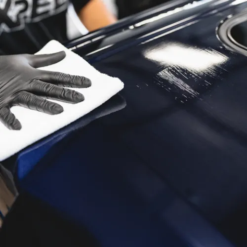 Person wearing black gloves polishing a glossy dark blue car surface with a white cloth.