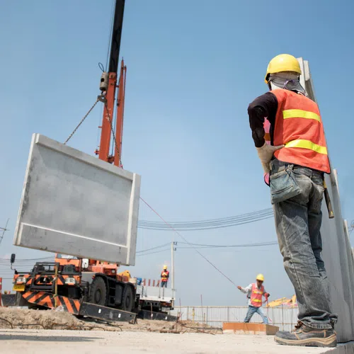 Construction workers wearing safety gear guiding a concrete panel being lifted by a crane at a building site.