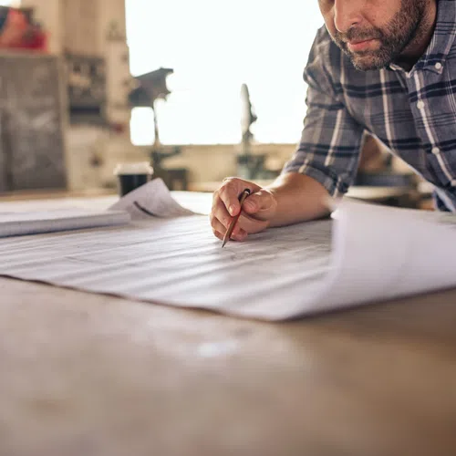 Man in plaid shirt reviewing architectural blueprints on wooden table in a workshop with natural light.