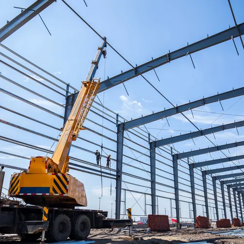 Mobile crane lifting steel beams during construction of large industrial warehouse under blue sky.