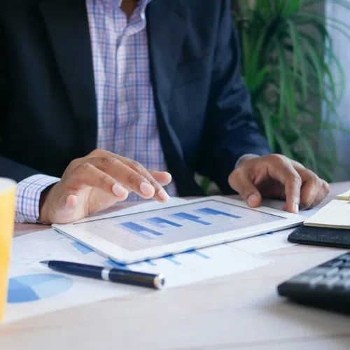 Businessperson analyzing bar charts on tablet with calculator, notebook, pen, and coffee cup on desk.