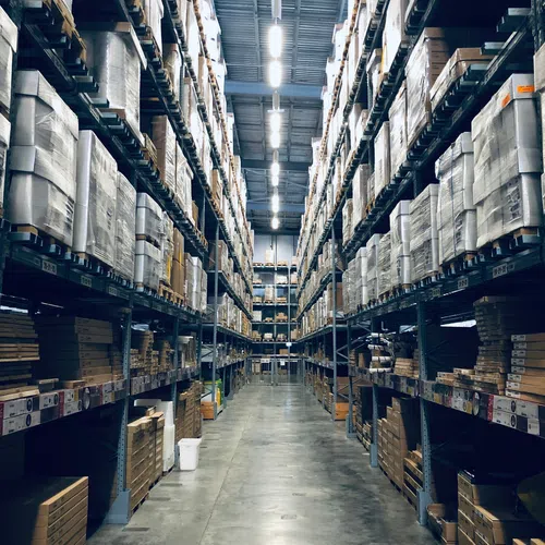 Long aisle in a large warehouse with tall shelves stocked with boxes and packages on both sides under bright lights