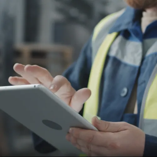 Construction worker in reflective vest using tablet device on job site with blurred background.