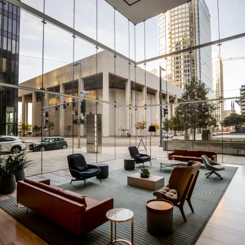 Modern lounge area with glass walls overlooking city street and tall buildings, featuring leather sofas and armchairs.