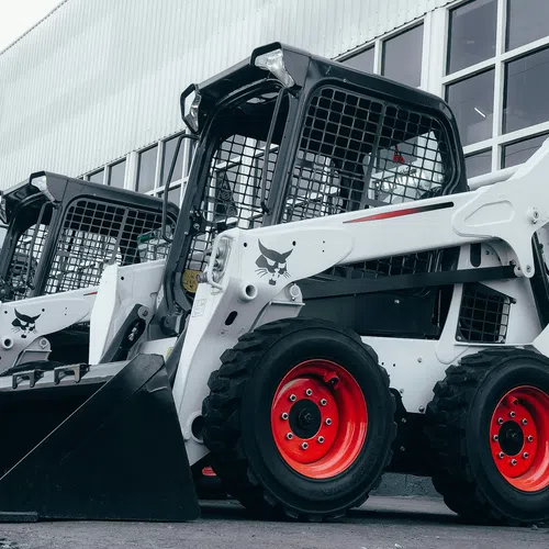 Row of white Bobcat S530 skid-steer loaders with red wheels parked outside a building under overcast sky