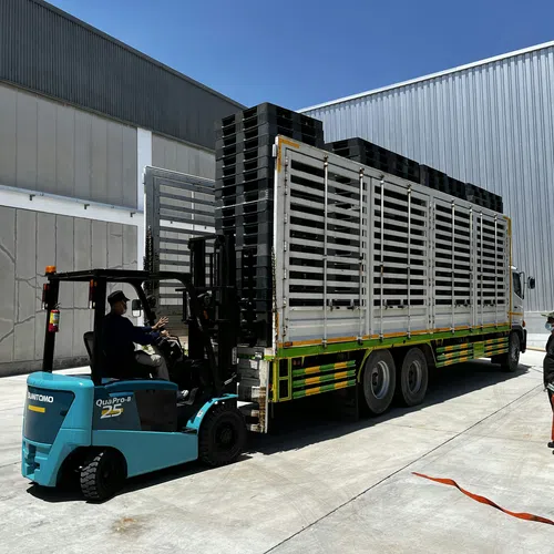 Forklift loading black crates onto a large white transport truck next to a warehouse under clear blue sky.