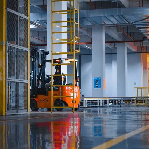 Orange forklift inside a spacious industrial warehouse with reflective polished floor and yellow safety railings.