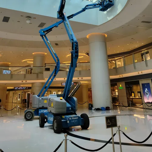 Blue articulated boom lift inside a shopping mall extending to clean or maintain the glass ceiling skylight.