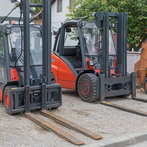 Two red and black forklifts parked side by side on gravel near a building with greenery in the background