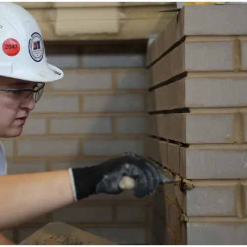 Woman wearing hard hat and gloves applies mortar to brick wall at construction site indoors.