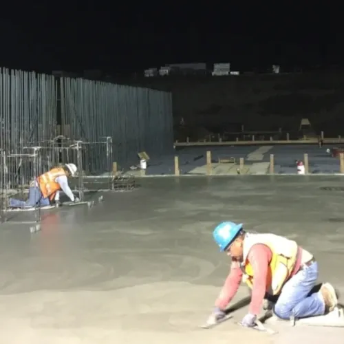 Construction workers smoothing freshly poured concrete slab at night on large building site under bright lights
