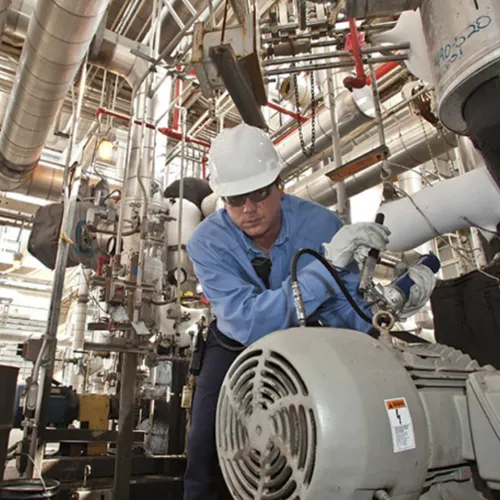 Industrial worker in blue uniform and hard hat inspecting machinery in a factory with pipes and equipment.