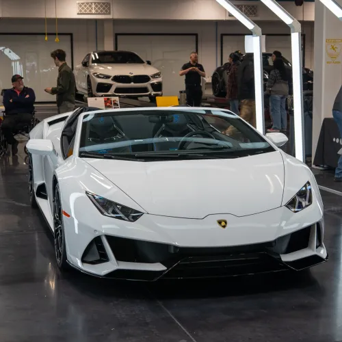White Lamborghini sports car displayed indoors with people and other cars in background under bright lights