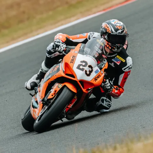 Motorcycle racer in full gear leaning into a sharp turn on an asphalt race track with orange and black bike number 23.