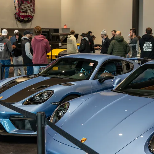 Two blue Porsche sports cars displayed indoors with people and a racing car backdrop in a showroom setting