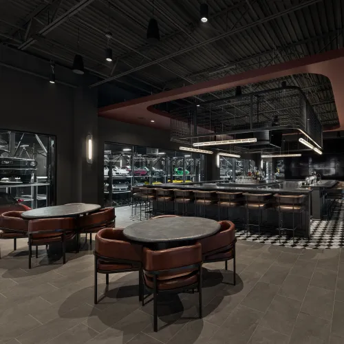 Modern bar area with leather chairs, round tables, and a luxury car display visible through glass walls.