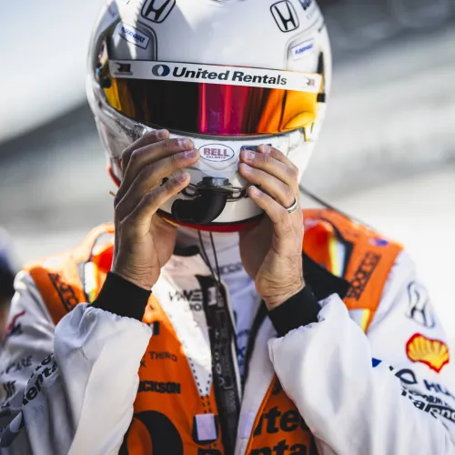 Race car driver adjusting white helmet with orange visor and United Rentals branding on race suit