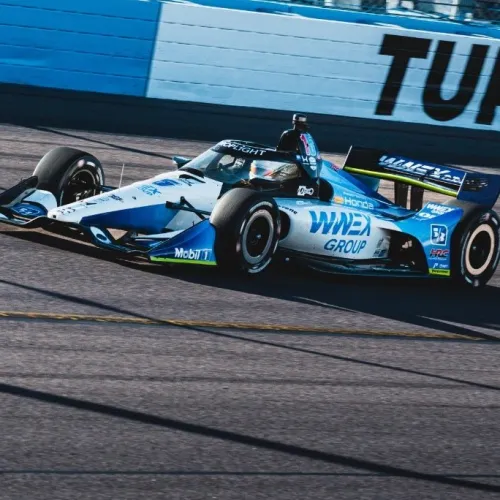 IndyCar race car speeding on race track at Turn 1 with tents and barrier walls in background