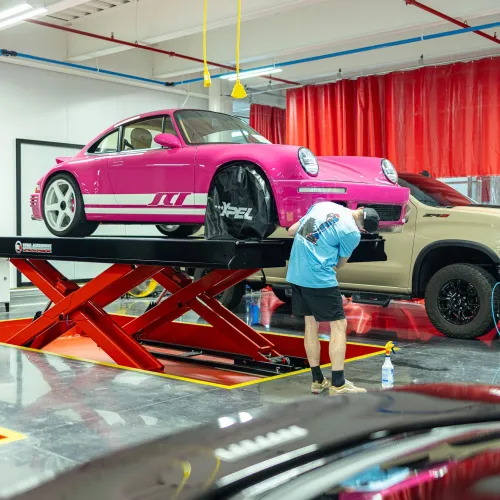 Man working on a pink classic car elevated on a red hydraulic lift in a modern auto workshop.